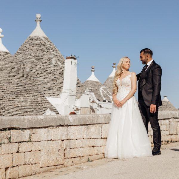 Portrait of happy wedding couple standing on street near conical roofs of trullo, meeting eyes. Young woman bride with long wavy fair hair wearing long white embroidered dress, hugging with groom.
